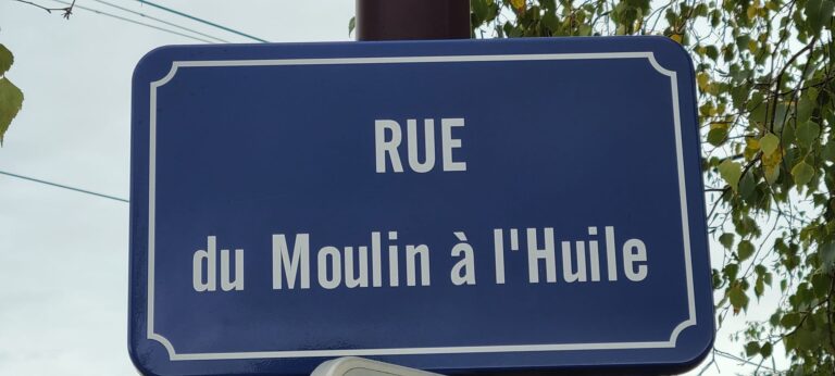 Plaque de rue en métal fixée sur un mur de granit gris, portant un nom gravé en lettres rouges, symbole de mémoire et de traces du passé.