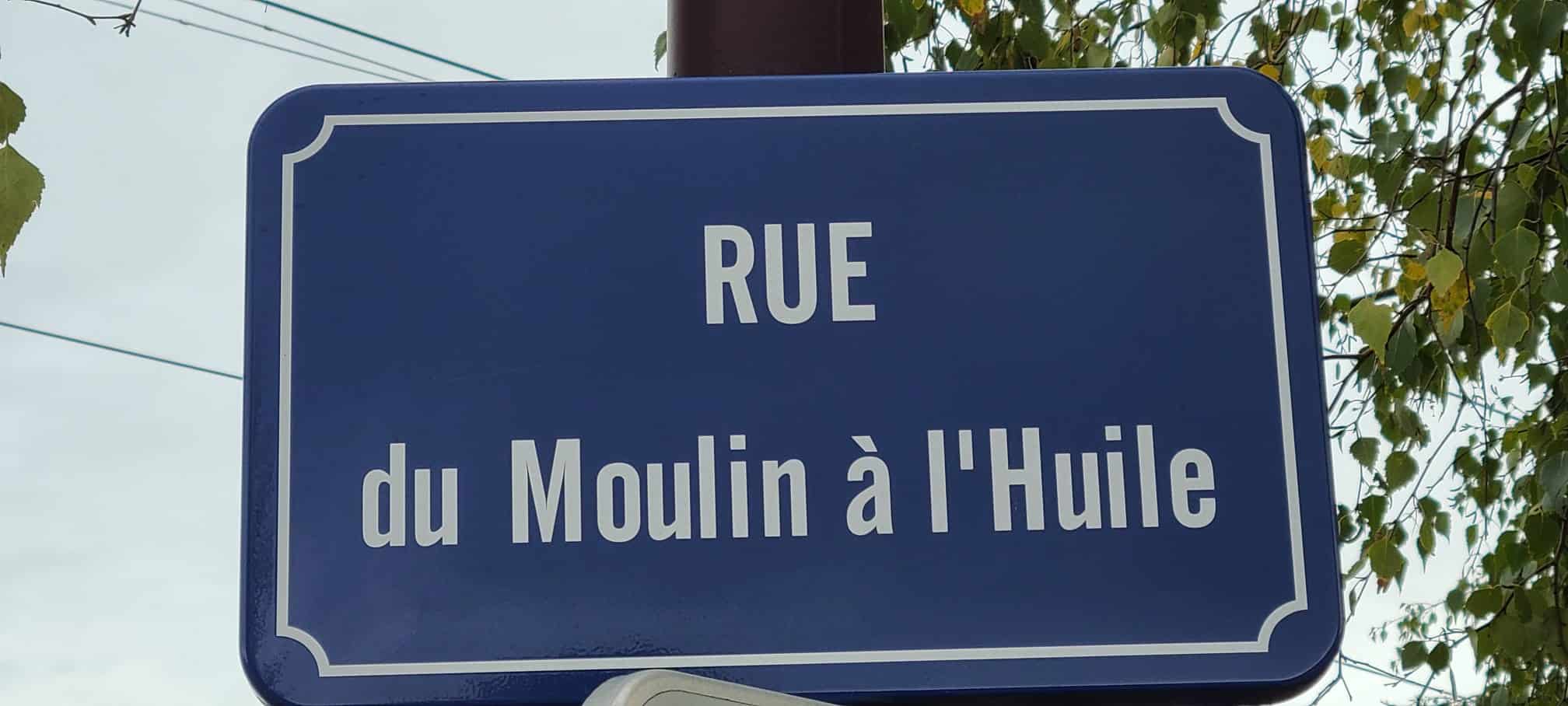 Plaque de rue en métal fixée sur un mur de granit gris, portant un nom gravé en lettres rouges, symbole de mémoire et de traces du passé.